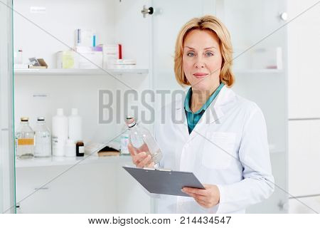 Moddle-aged nurse or assistant with bottle of medicine and clipboard standing by cabinet with glassware