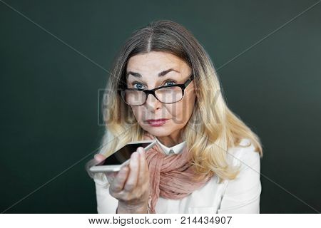 Charming elderly woman in rectangular black rimmed eyewear using voice recognition function on cell phone. Modern senior female holding electronic gadget sending vocal message via online messenger