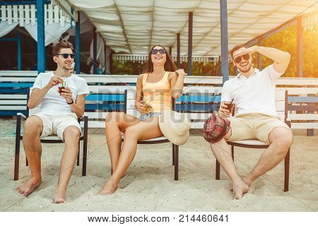 Lifestyle Young people enjoying summer vacation sunbathing drinking at beach bar. Two guys and a young girl sit on the terrace of the summer bar and enjoy the conversation Sun flare