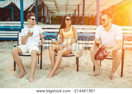 Lifestyle Young people enjoying summer vacation sunbathing drinking at beach bar. Two guys and a young girl sit on the terrace of the summer bar and enjoy the conversation Sun flare