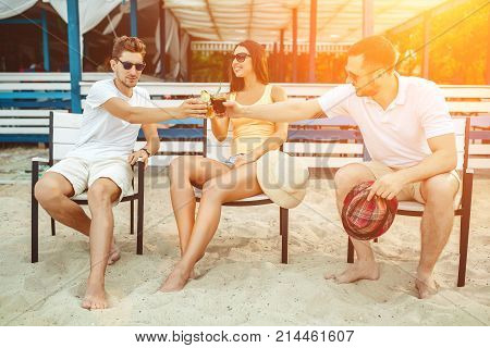 Lifestyle Young people enjoying summer vacation sunbathing drinking at beach bar. Two guys and a young girl sit on the terrace of the summer bar and enjoy the conversation Sun flare