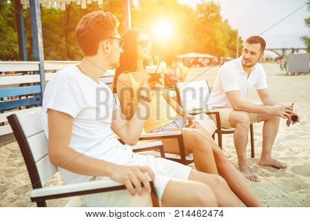 Lifestyle Young people enjoying summer vacation sunbathing drinking at beach bar. Two guys and a young girl sit on the terrace of the summer bar and enjoy the conversation Sun flare