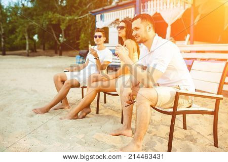 Lifestyle Young people enjoying summer vacation sunbathing drinking at beach bar. Two guys and a young girl sit on the terrace of the summer bar and enjoy the conversation Sun flare
