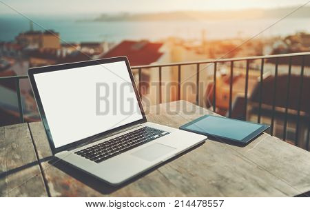 Modern laptop with blank screen mockup and huge trackpad on the wooden table next to tablet pc; netbook with empty white screen template near digital tablet on balcony workspace with blurred cityscape