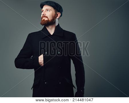 Portrait of Redbeard elegant male dressed in a tweed flat cap and a black jacket over grey background.