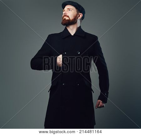 Portrait of Redbeard elegant male dressed in a tweed flat cap and a black jacket over grey background.