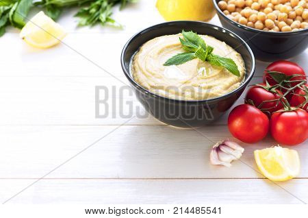 Bowl with hummus, chickpeas, lemon, cherry tomatoes and herbs on a white rustic wooden background.
Middle Eastern cuisine. Background hummus with place for text.
