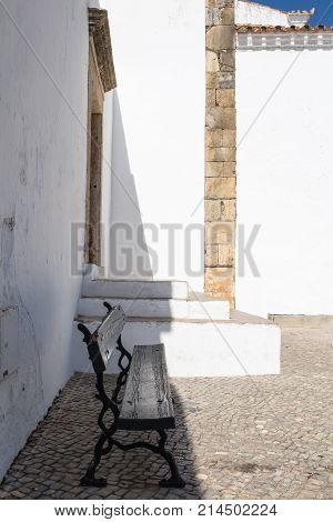 Wooden bench on a street with cubble stones. Sunny day bench remaining in a shadow of a white old building. Faro Portugal