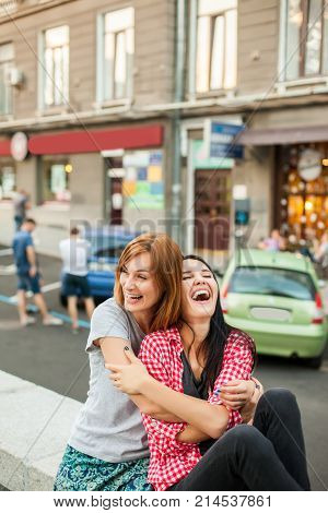 Two young girls hugging, laughting and sitting near road in the street. one brunette girl in red plaid shirt, another redhead girl wearing gray shirt and blue skirt. concept of sincere friendship