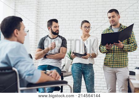 A man in a wheelchair is showing documents to his team in office. They are showing a teamwork. They discus something realy important.