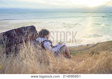 Boy reading book outside on the top of the hill, snacking with healthy apple, unschooling education