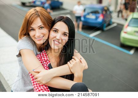 Two atractive young girlfriends hugging, laughting and sitting near road in the street. one girl in red plaid shirt, another redhead girl in gray shirt and blue skirt. concept of sincere friendship