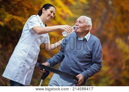 Medical professional helping a senior man with walker drink a wather in park