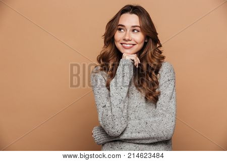 Young attractive woman in woolen sweater holding her chin, looking aside, isolated on beige background