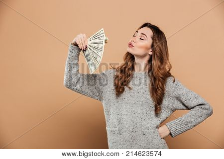 Portrait of gorgeous brunnete woman with closed eyes waving with fan of money, isolated over beige background