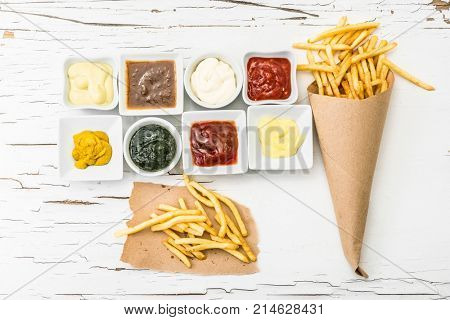 French fries on a piece of crafted paper with different sauces in white sausers on the white wooden background with cracking paint