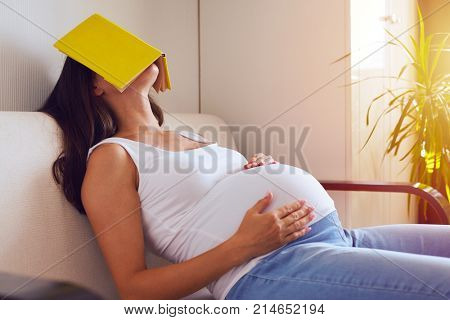 Side view of pregnant woman tired of reading, sleeping on sofa with a book on her face
