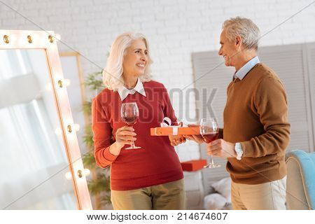 Happy anniversary darling. Joyful elderly couple exchanging gift and drinking red wine while celebrating their anniversary at home.
