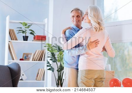 Romantic mood. Positive minded retired couple looking into vacancy with broad smiles on their faces while both dancing at home.