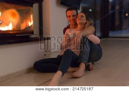Young romantic couple sitting on the floor in front of fireplace,talking and drinking tea on cold winter night at home