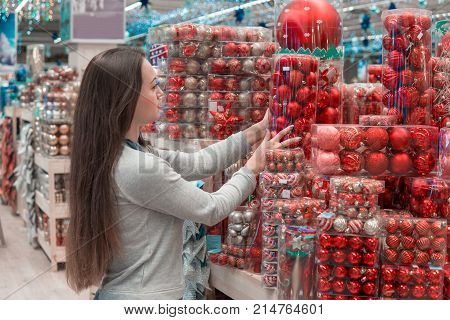 Cheerful happy customer girl choosing gifts for Christmas and New Year. She is taking red Christmas tree decorations. difficult decision. Various choice