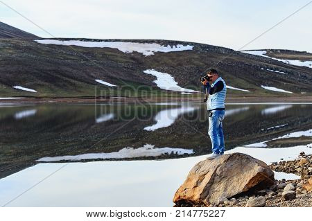 Horizontal shot of landscape photographer standing on stone and wearing blue jeans and pale blue vest photographing mountain lake with melting snow