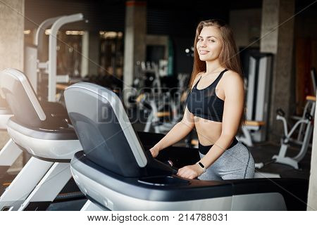 Portrait of crazy fit woman on a threadmill smiling looking at camera. Athletic coach warming up early in the morning in empty gym.