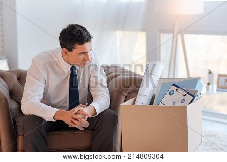 Hardships. Thoughtful serious young man looking at the box with documents and graphics while sitting in a brown armchair and looking at the box with documents after being dismissed