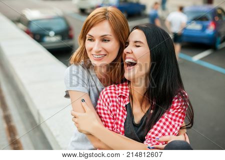 Two young sisters hugging, laughting and sitting near road in the street. one brunette girl in red plaid shirt, another redhead girl wearing gray shirt and blue skirt. concept of sincere friendship