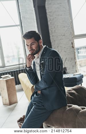 Thinking about something pleasant. Handsome young man in full suit smiling and keeping hand on chin while leaning on the sofa