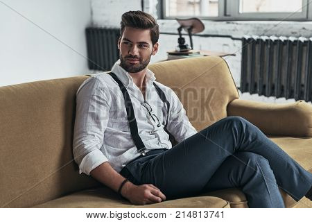 He knows all of your secrets. Handsome young man in white shirt and suspenders looking away and smiling while sitting on sofa