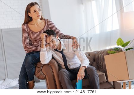Reliable person. Depressed exhausted man sitting in an armchair and feeling guilty after telling his young loving wife about his unemployment