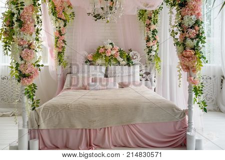 Honeymoon suite with canopy bed free space. Luxurious wood canopy bed with flowers and pillows on it. Female bedroom in pink and white colors copy space. Big comfortable bed in elegant bedroom