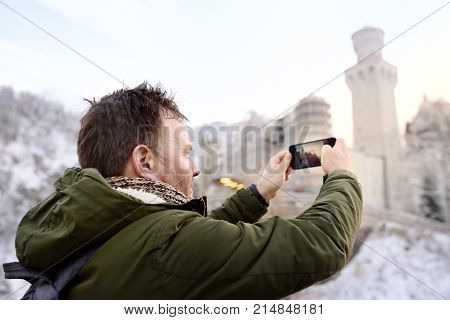 Handsome milddle age man making photo of famous royal castle Neuschwanstein. Happy winter weekend in the countryside. Vacation in mountains. 
