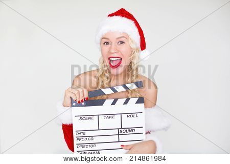 Portrait of cheerful young Caucasian woman wearing Santa costume holding clapperboard and looking at camera with open mouth. Christmas and Film production concept