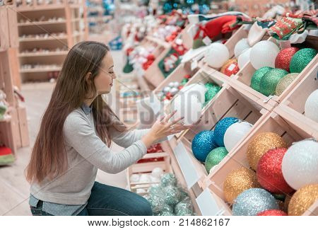 Cheerful happy customer girl choosing gifts for Christmas and New Year. She is taking red Christmas tree decoration balls. difficult decision. Various choice
