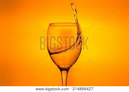 Wineglass with splashing drops of fresh water - motion freeze close-up picture isolated on the orange background.