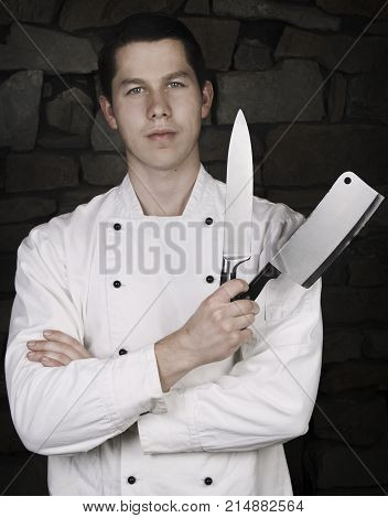 Illuminated portrait of nice and attractive man chef in uniform on the stone wall background.  Man chef posing to camera with knife and cleaver (chopper) knife on dark background.