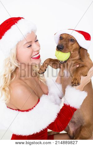 Closeup portrait of smiling young attractive woman wearing Santa Claus hat and holding dog which is holding ball. Isolated side view on white background.