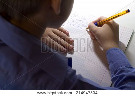 Close-up of boy hand with pencil writing english words by hand on traditional white notepad paper. Boy writes a letter to a friend