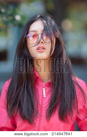 Asian woman fashion portrait. Sensual seductive lovely and beautiful mixed race asian caucasian young girl in pink jaket and red sunglasses looking at camera outdoor against green blurred bokeh city background. Gorgeous slim model posing outside on the st