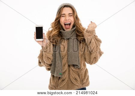 Screaming ecxited young woman dressed in warm sweater and scarf standing isolated over white wall background showing display of mobile phone.
