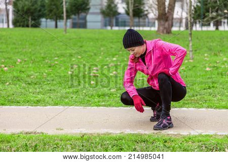 Young woman exhausted after train on a cold winter day on the training track of an urban park. Female athlete wearing pink windbreaker, beanie, gloves and running tights