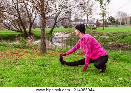 Young woman warming up and stretching the legs before running on a cold winter, autumn of fall day in an urban park. Female athlete wearing pink windbreaker, beanie, gloves and running tights