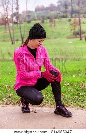 Female athlete hurting from a knee injury on a cold winter day on the training track of an urban park. Young woman wearing pink windbreaker, beanie, gloves and running tights