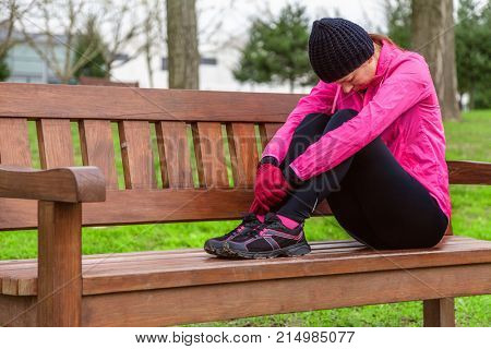 Female athlete tired or depressed resting on a bench on a cold winter day on the training track of an urban park. Young woman wearing pink windbreaker, beanie, gloves and running tights