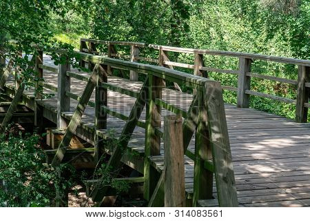 Scenic Wooden Pedestrian Bridge Over A River With Wild Trees And Plants In The Background