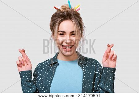Wishful Young Woman Smiling Broadly, Raises Fingers Crossed, Makes Desirable Wish, Wears Casual Outf