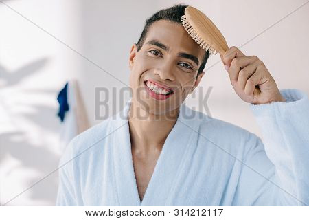 Handsome Young Man In Bathrobe Combing Hair With Hairbrush While Amiling And Looking At Camera