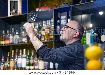 Elderly Bartender Cleans The Glass. A Handsome Bartender Polishes A Glass Of Wine Glasses. The Conce